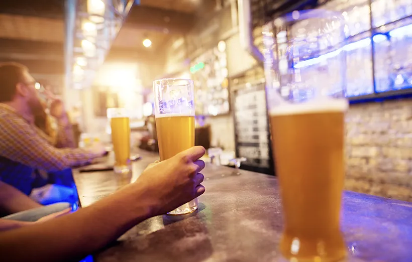 Close up view of hand while holding draft beer glass placed on a bar table with a row of other in the background.