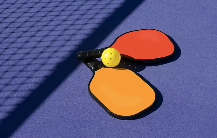 Simple, colorful composition of pickleball paddles and pickleball lying on court near the shadow of the net.