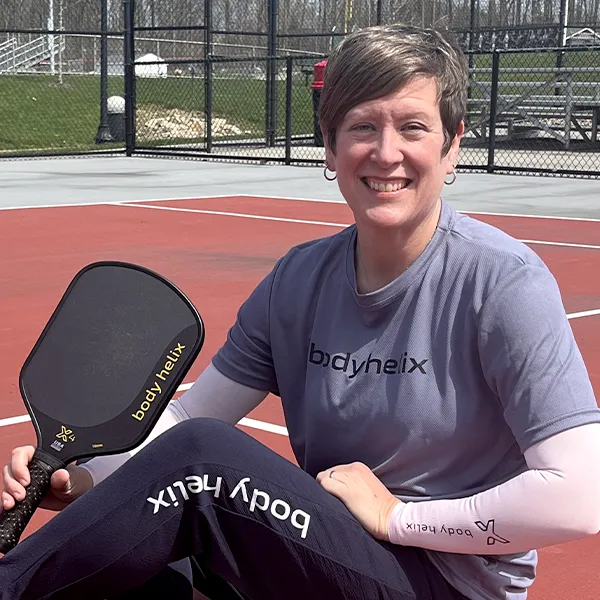 Portrait of Tracey Flynn sitting on pickleball court holding a racket.